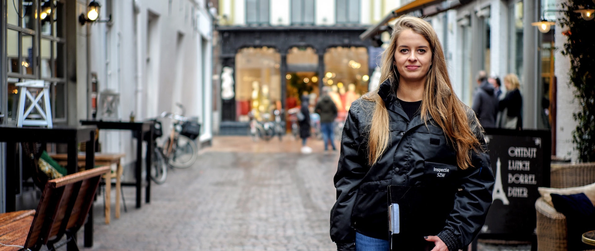 Vrouwelijke inspecteur in jas van Nederlandse Arbeidsinspectie en met notitieblok in een winkelstraat
